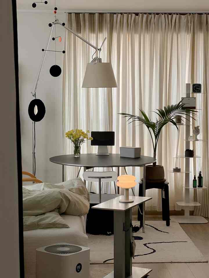 ベージュと白のトーンのワンルーム玄関スペース、カーテン、丸テーブル、植物、照明が配置された雰囲気のあるインテリア