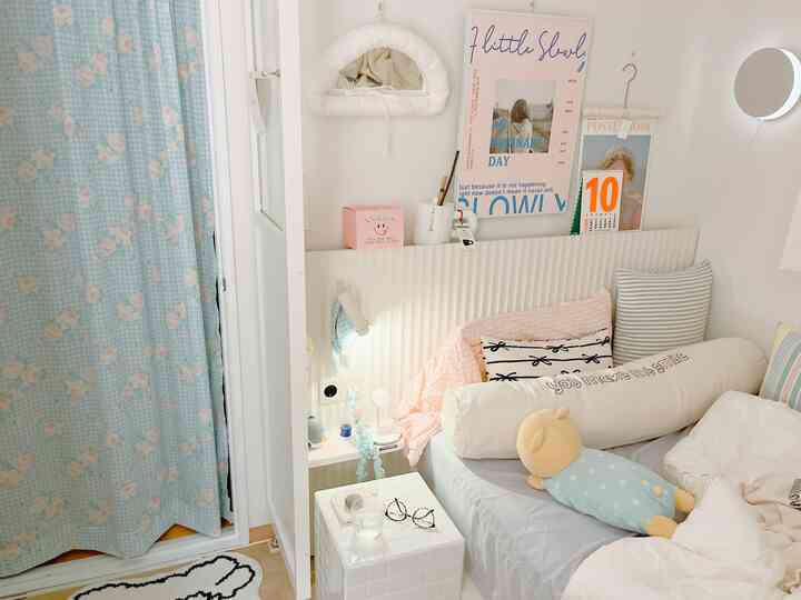 ホワイトとブルーの色調の狭く縦長の寝室、ベッドの横に花柄のカーテンと複数のクッションが配置された雰囲気のあるインテリア