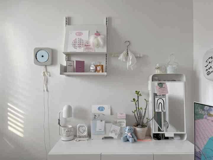 白を基調としたシンプルで整ったデスクスペース、壁掛け棚と時計、植物、テーブルランプが配置された雰囲気のあるインテリア