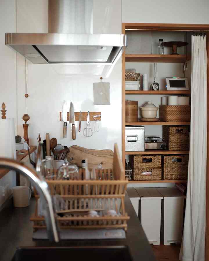 白い壁とナチュラルなブラウンの木製棚があるキッチンのパントリー、リネンカーテンと収納バスケットが配置された清潔感のある収納スペース