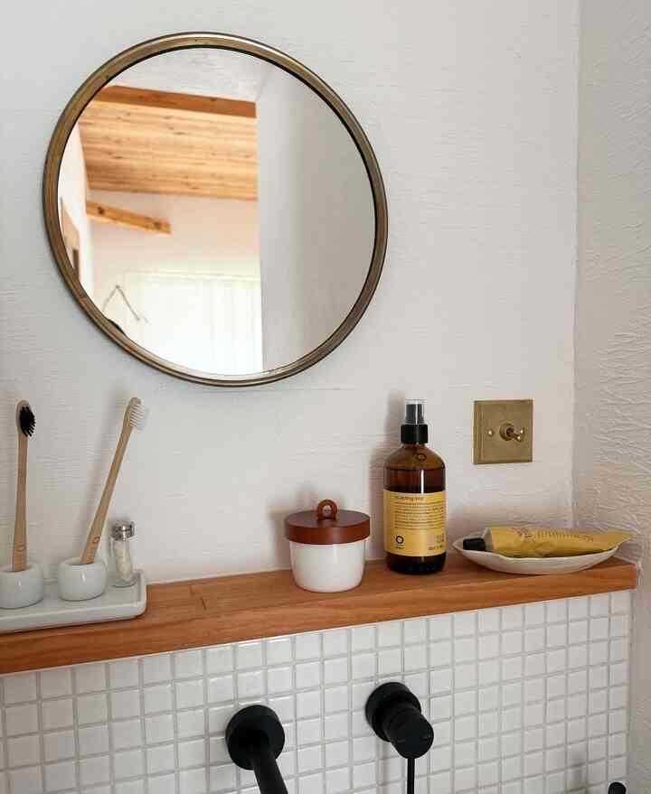 白いタイルと木目調の棚があるバスルーム、丸い鏡と濃い茶色のボトル、真鍮スイッチが配置された清潔感のある空間