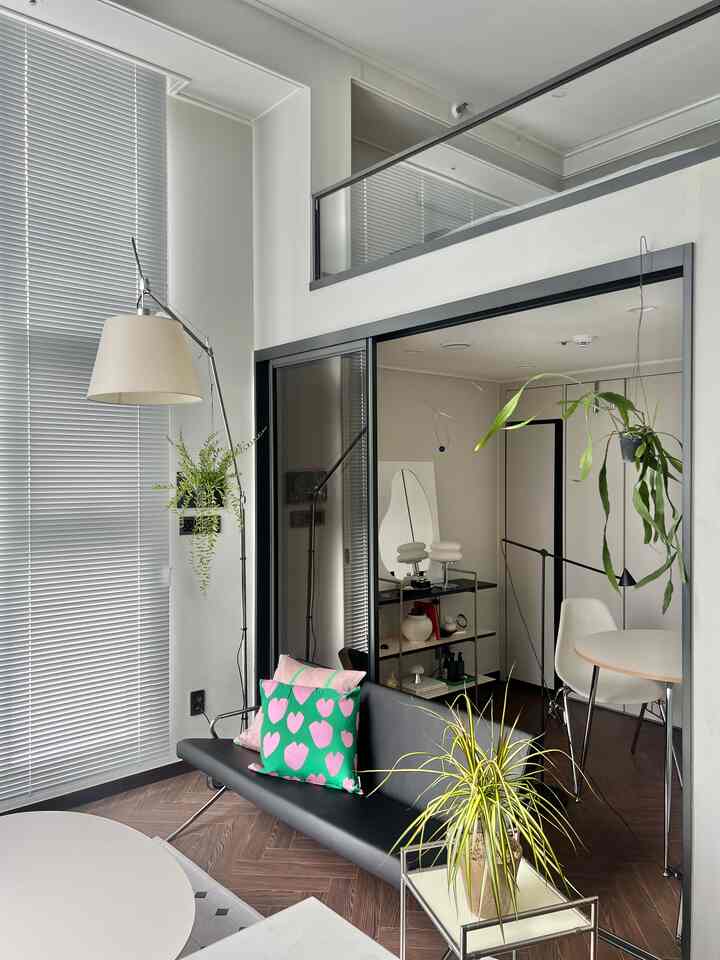 ホワイトの壁とブラインドが印象的な広く見えるリビング、ブラックのソファとグリーンクッション、植物が調和するモダンな空間