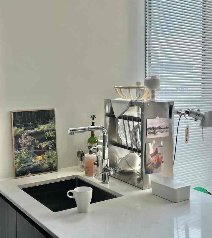 白を基調としたキッチン、シンクに白いマグカップ、シルバーの食器棚、額縁が配置された清潔感のあるインテリア