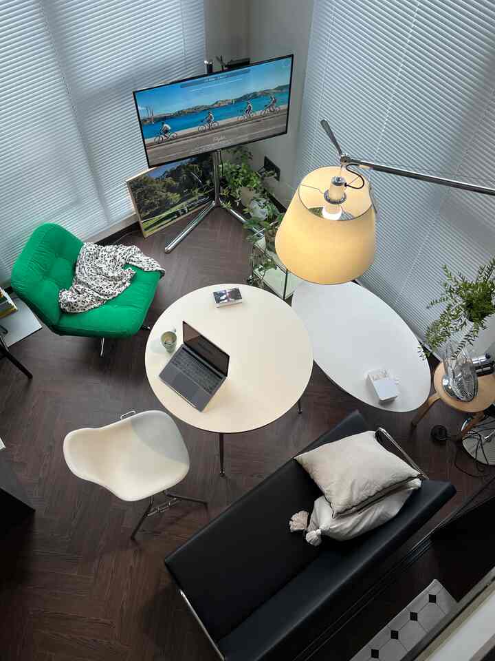 ナチュラルトーンの円形の部屋にモダンな家具、ノートパソコンと大型モニターを備えた作業スペース、グリーンのアームチェアが配置された雰囲気のあるホームオフィス