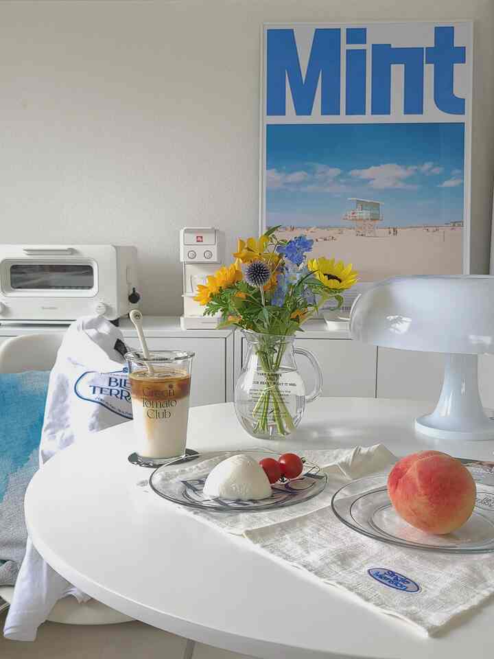 白を基調としたキッチン兼カフェ風ダイニングスペース、中央には花瓶と飲み物、食事がセットされた丸テーブルが配置された清潔感のある空間