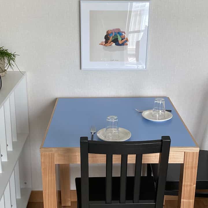 青い天板の木製脚ダイニングテーブルと黒い椅子が配置されたシンプルなダイニング、壁には家族の絵の額縁が飾られている