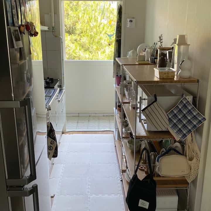 明るいベランダの窓がある狭い通路型キッチン、棚と冷蔵庫が配置された実用的な一人暮らしの空間