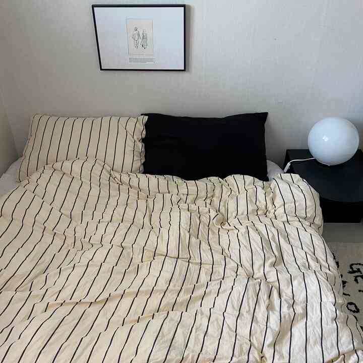 ベージュのストライプ柄掛け布団カバーと黒い枕が配されたシンプルでモダンな寝室、壁には額縁が飾られ、ナイトテーブルの上には白い球形の照明が置かれている