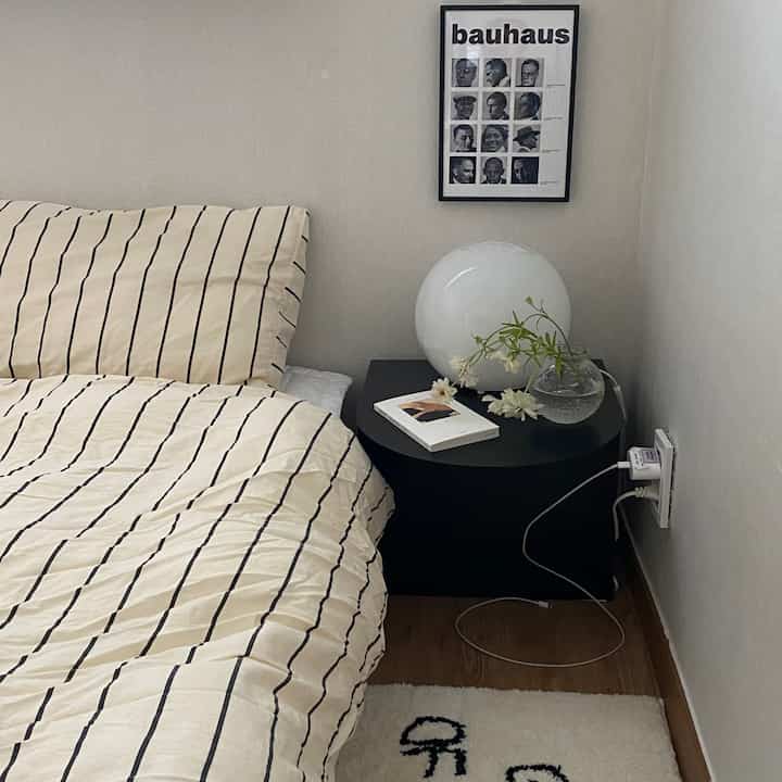 ベージュとブラックの色調の寝室、ストライプの寝具と黒いナイトテーブル、花瓶と丸い照明が配置された雰囲気のあるインテリア