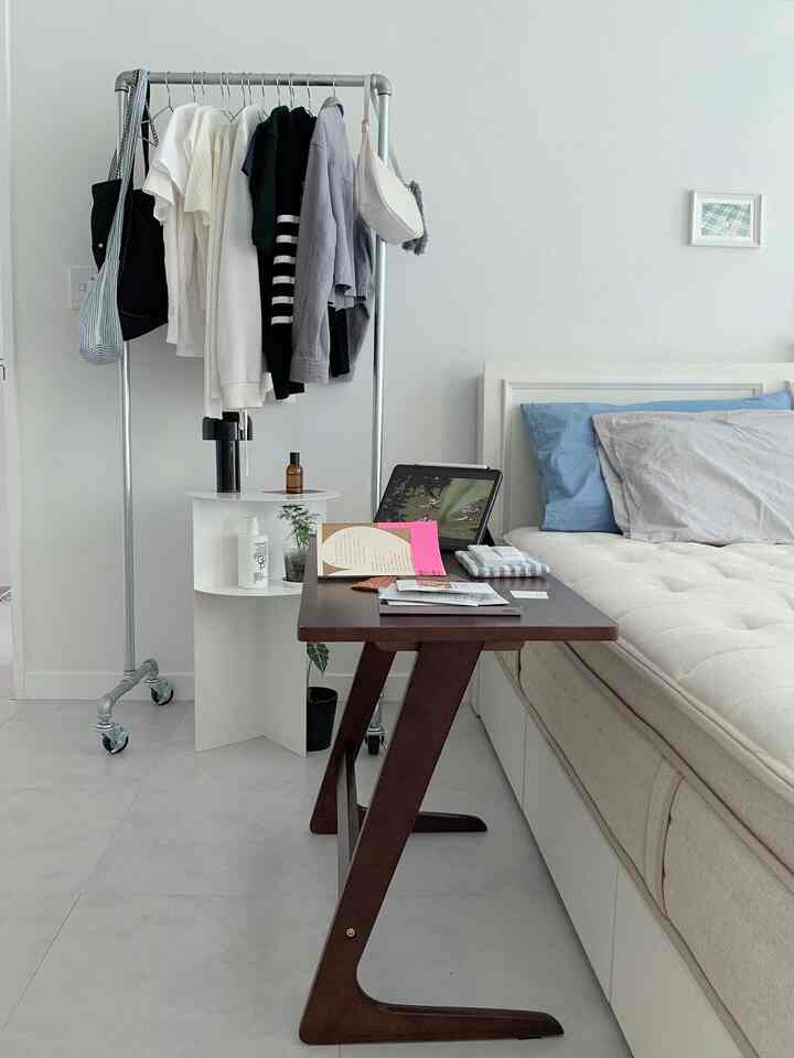 ホワイトとブラウンのトーンが調和した寝室、ベッド、サイドテーブル、移動式ハンガーラックが配置されたすっきりとしたインテリア