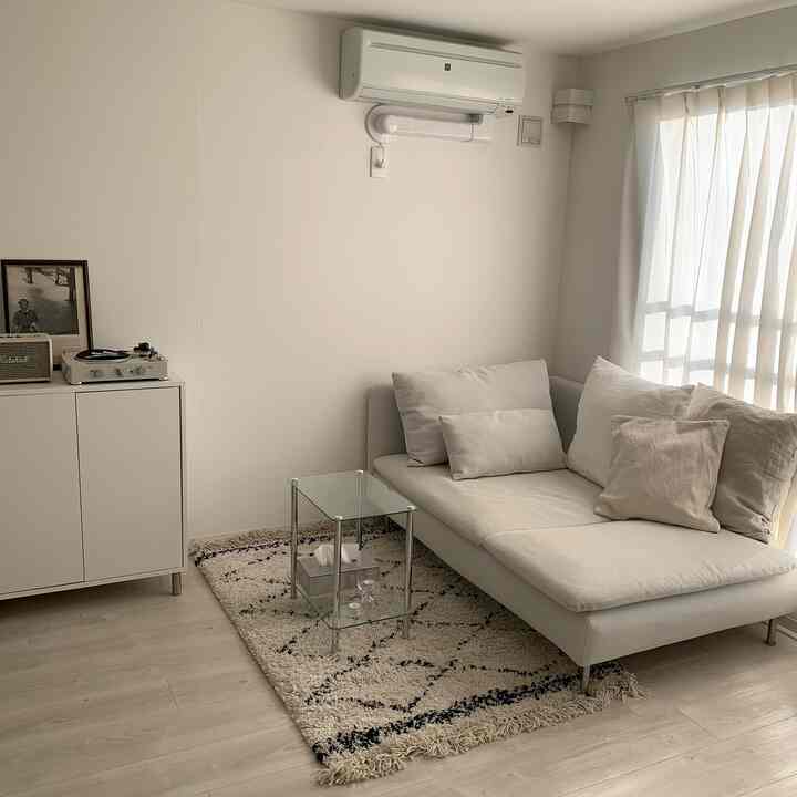 ナチュラルカラーのコンパクトなリビングルーム、ベージュのソファとガラスのサイドテーブル、白いカーテンと壁が配置されたシンプルモダンな雰囲気のあるインテリア