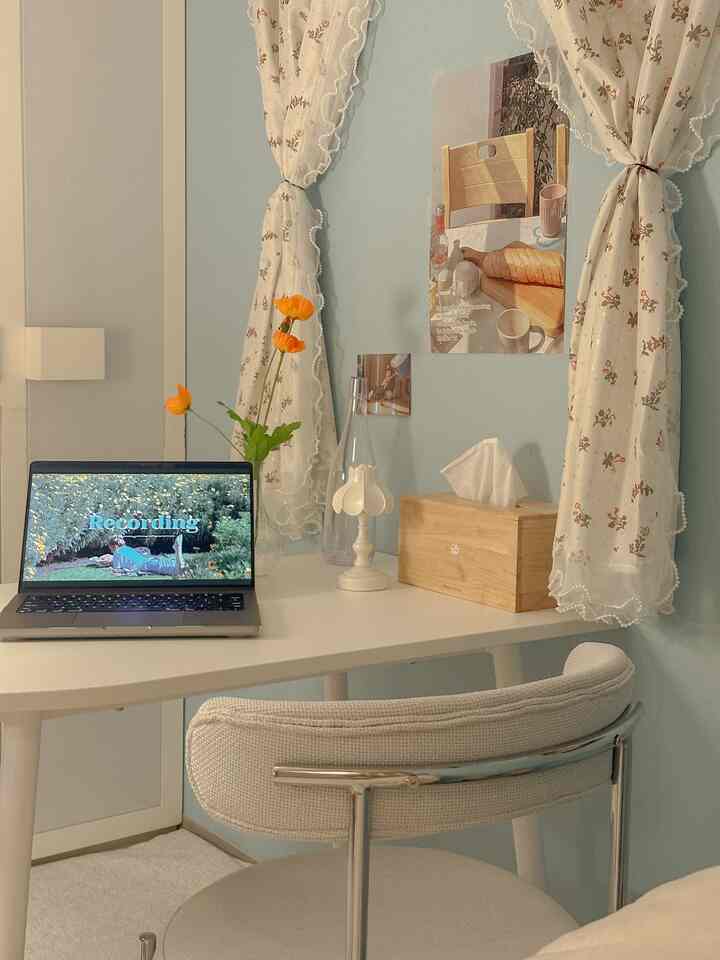 ホワイトとライトブルーの色調の狭いホームオフィス空間、ホワイトのデスクとベージュの椅子、繊細なテーブルランプとポスターが配置された落ち着いた印象のインテリア