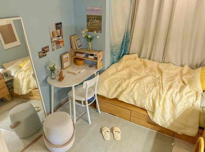 パステルブルーの壁と木目調ベッドがあるコンパクトな一人用寝室、シンプルな白いデスクとクリーム色の寝具が配置された空間