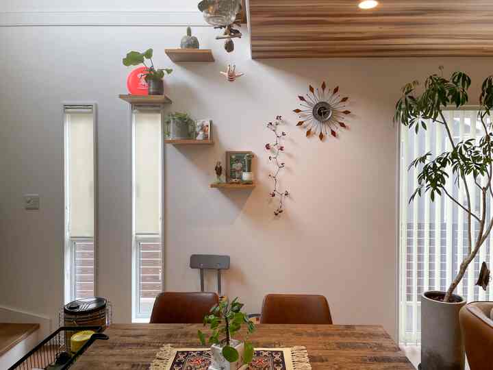 白い壁と木製の天井が調和するダイニング、木製のテーブルと茶色の椅子、壁棚と時計、そして植物が配置された自然な雰囲気のインテリア