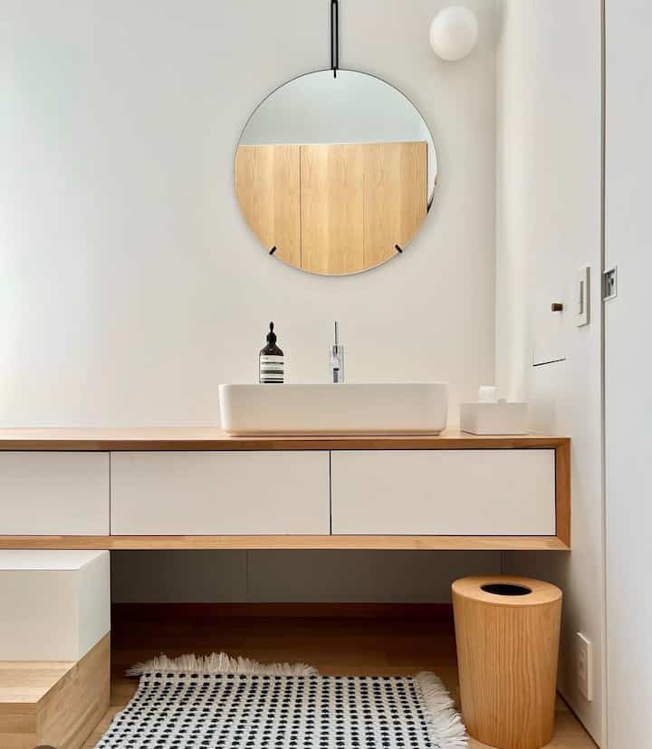 ナチュラルな色調のバスルーム、丸い鏡と木製フレームの洗面台が配置された清潔感のある空間