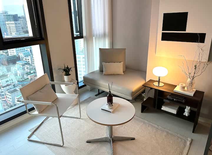 木目調と明るいニュートラルカラーが調和する小さなリビング、大きな窓とアームチェア、ラウンジソファが暖かい雰囲気を演出しているインテリア