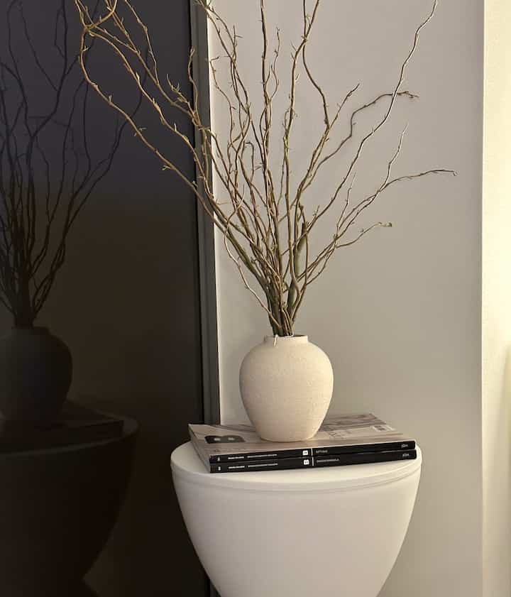 白とナチュラルトーンの空間、スツール上に配置されたカーリーウィローブランチとテクスチャードベースがあるミニマルなインテリア