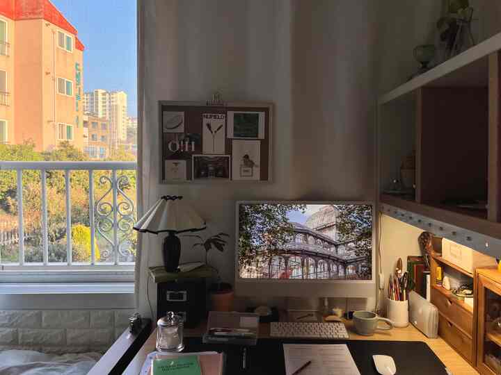 白と木目調のワンルームホームオフィス、デスクやパソコン、テーブルランプ、植物とバルコニーの景色があり、落ち着いた雰囲気のインテリア。