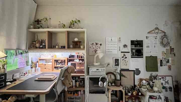 白い壁と木目調家具が調和するコンパクトなホームオフィス空間、デスク上のパソコンと植物、ポスターや多様な小物が壁に配置された雰囲気のあるインテリア