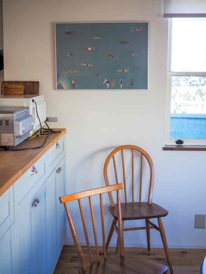 白い壁とナチュラルな木目調の椅子が配置された静かなキッチン、中央上部に泳ぐ人々のポスターが飾られている印象的な空間