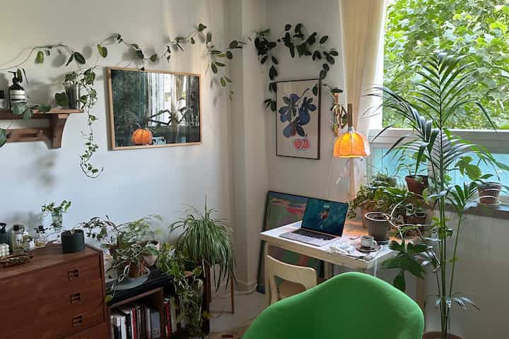 ナチュラルトーンのリビング兼ホームオフィス、豊かなグリーンの植物とヴィンテージ家具、温かみのある照明が調和した印象的な空間