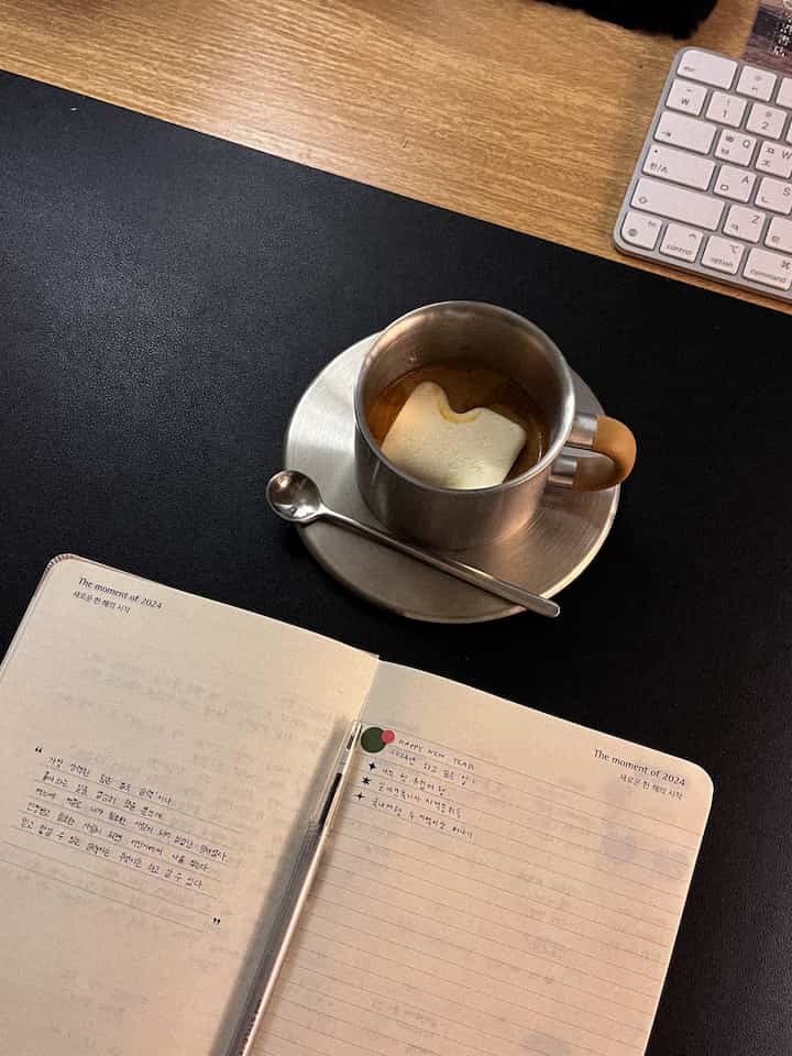 木目調のデスクに黒のデスクマット、アップルマジックキーボード、マシュマロ入りのコーヒーが入ったステンレスカップ、ノートとペンが置かれた落ち着いたおうちカフェの雰囲気のあるインテリア