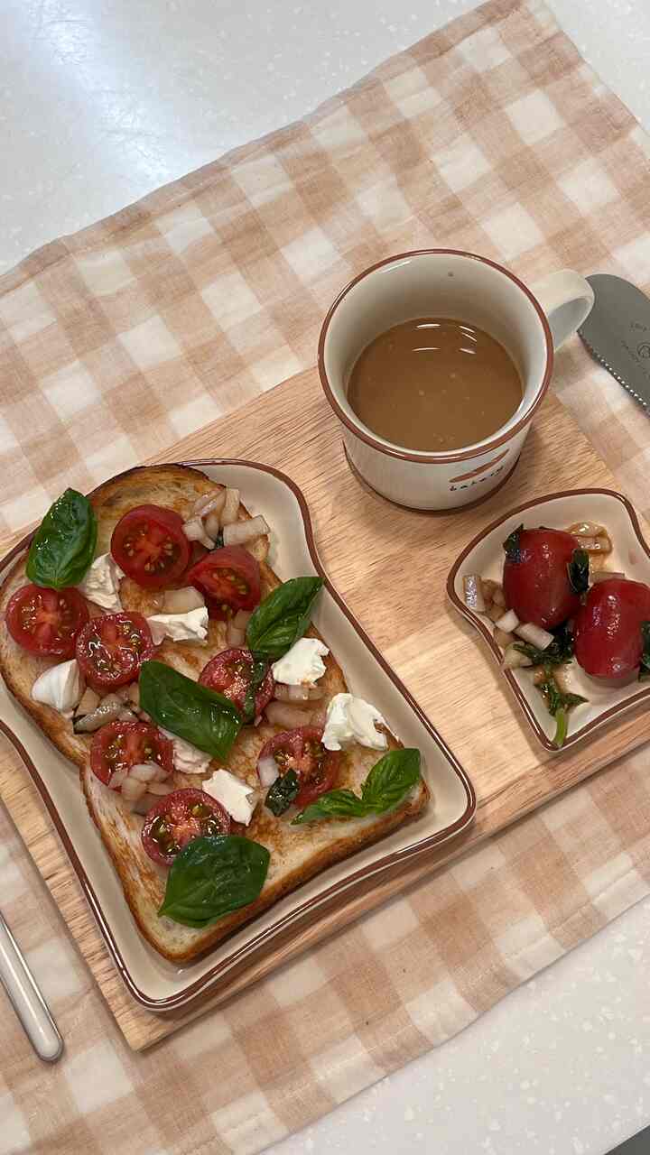 ベージュのチェック柄テーブルクロスの上に、トマト、バジル、クリームチーズをのせたトーストとオレンジ縁のマグカップが置かれたおうちカフェ風のテーブルセッティング