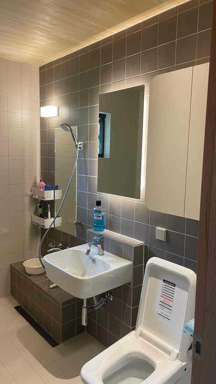 ナチュラルな木目調天井と茶色のタイル壁があるバスルーム、白い洗面台とトイレ、LED照明付きの鏡が配置されたモダンな空間