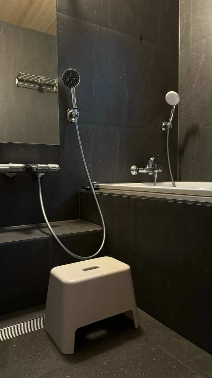 ダークブラックタイル仕上げのモダンなバスルーム、浴槽とシャワー、白いバススツールが配置された空間