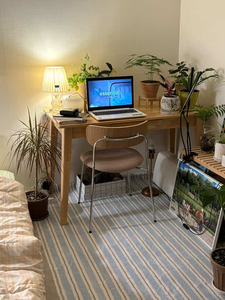 ナチュラルな色調の小さなホームオフィス、木製のデスクと複数の植物、ノートパソコンとテーブルランプが配された雰囲気のある空間