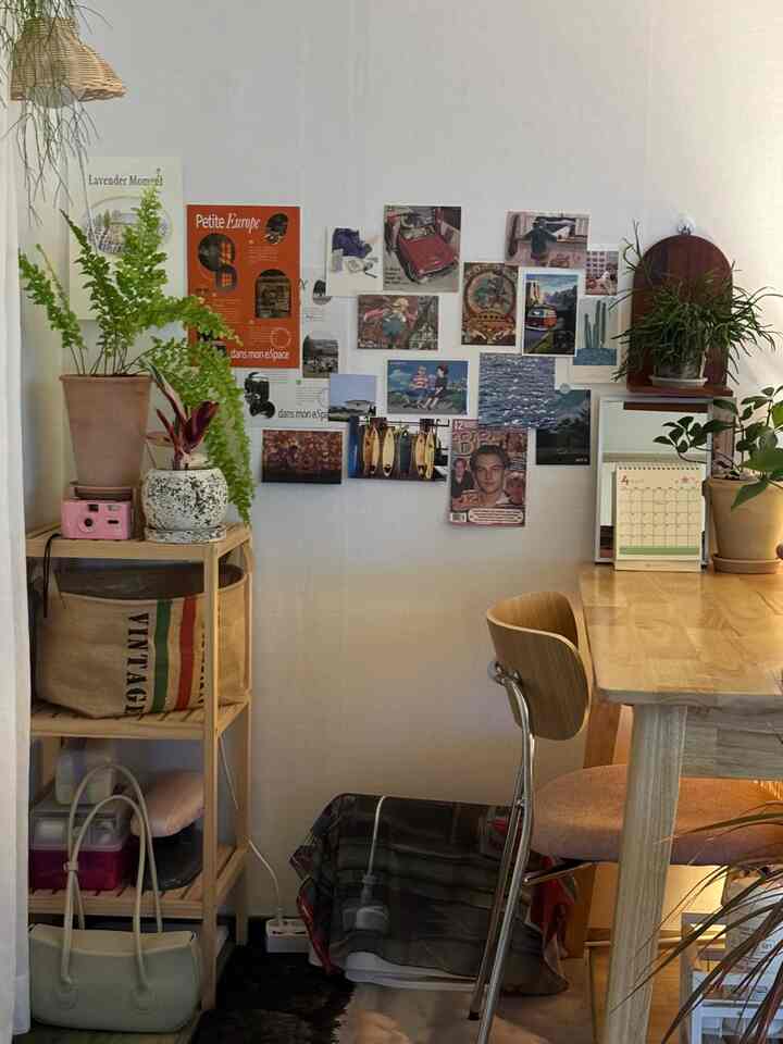 白い壁とブラウンの木目調家具が調和した書斎、机横に植物とヴィンテージポスターが配置された落ち着く空間