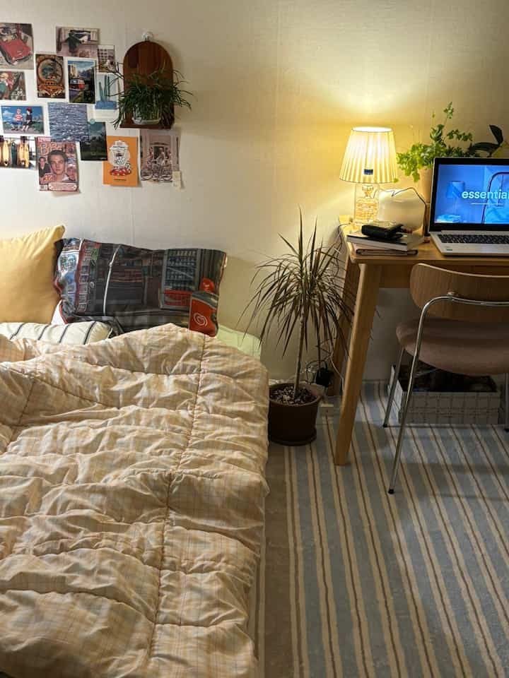 ベージュトーンの壁と寝具、木目調のデスクとラグが調和した一人暮らしの温かみのある寝室空間