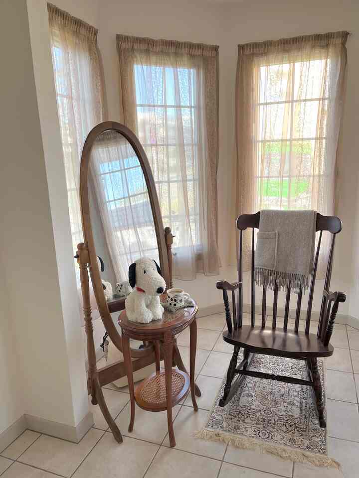 ベージュトーンのカーテンとタイル床がある玄関スペース、木製のロッキングチェアとサイドテーブル、そして大きな楕円形の鏡が配置された印象的な空間