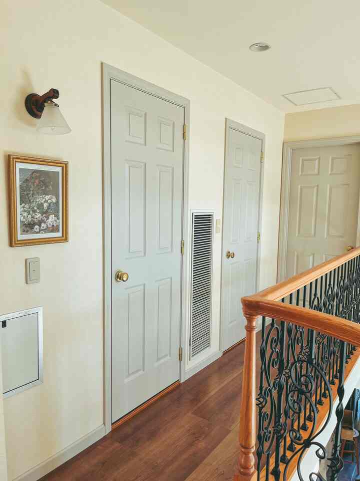 ベージュ調の壁とダークブラウンの木床を持つ2階の廊下、クラシックな扉と壁付け照明、木製手すりが配置された雰囲気のある空間