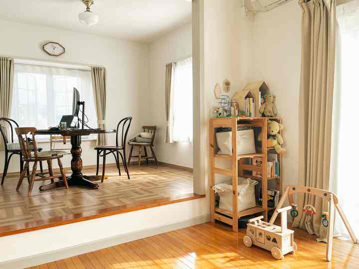 温かみのある木目調とベージュのカーテンがあるリビング、子供のおもちゃと収納ラックが配置された居心地のよい空間