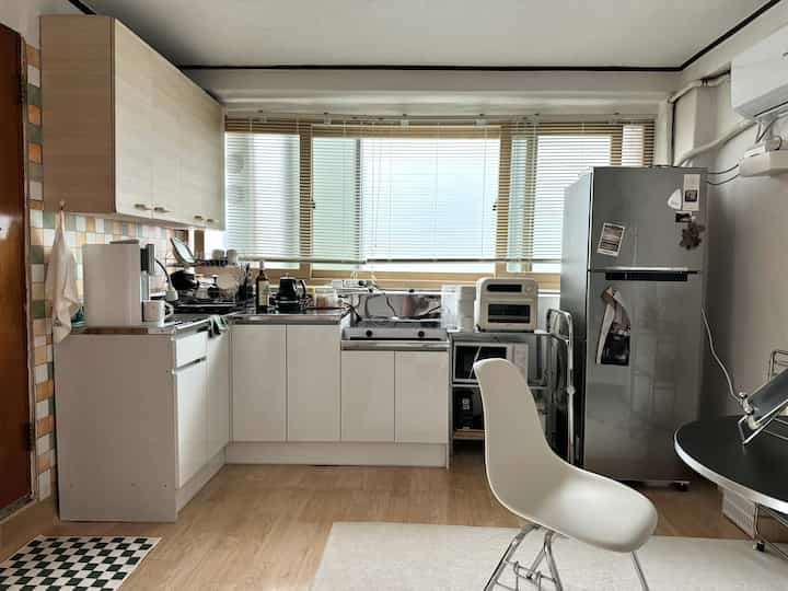 ホワイトと木目調の調和が美しいコンパクトキッチン、ミッドセンチュリーモダンの椅子と冷蔵庫が配置された実用的な空間