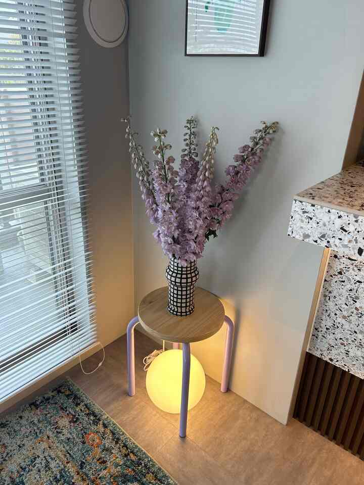 白い壁とブラインド、ベージュの床が調和するコーナースペースに、ラベンダーの花と間接照明を備えた木製スツールが配置された印象的な空間