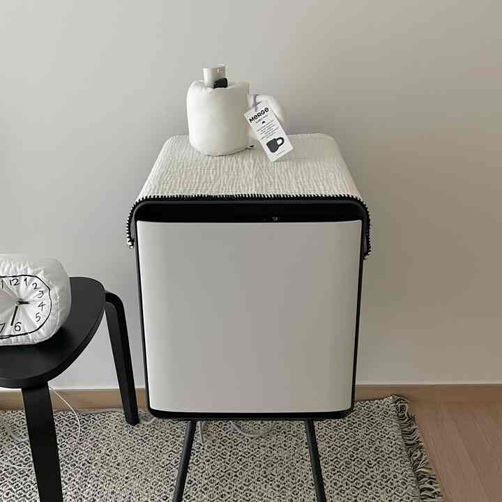 明るい白色とナチュラルな色調の寝室、中央に白いスツールとパターンラグ、黒いテーブルと時計が配置された清潔感のあるインテリア