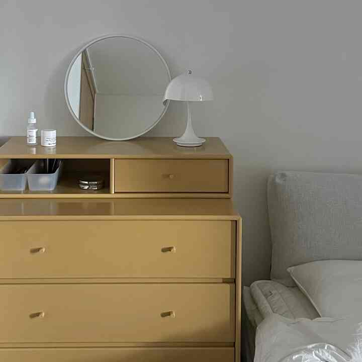 白い壁を背景に、黄色がかったベージュのチェストが置かれ、上に白いテーブルランプが配された寝室のインテリア