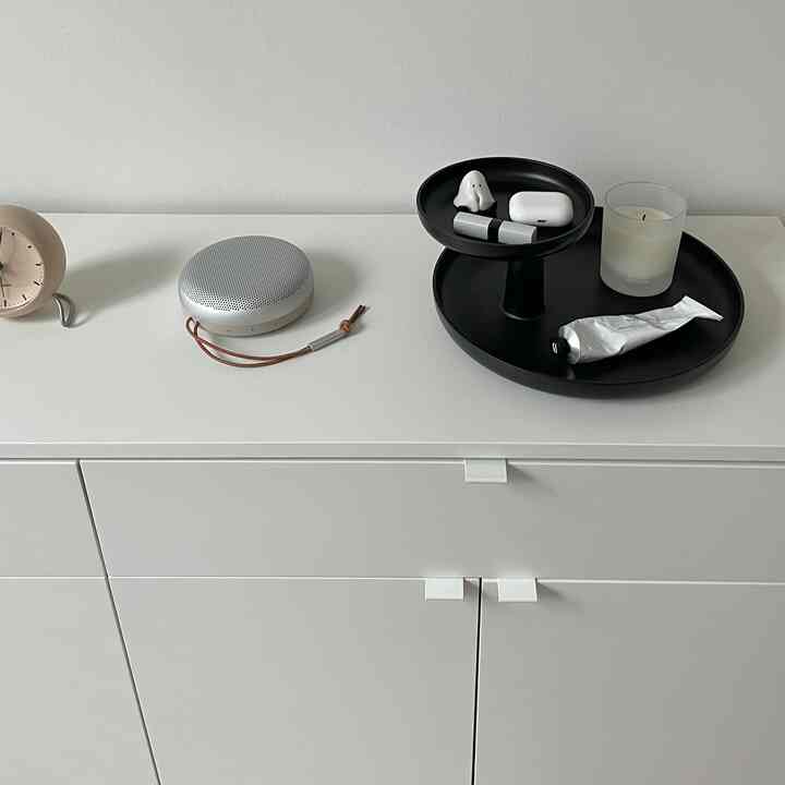 白を基調とした収納キャビネットの上に時計、Bluetoothスピーカー、黒いトレーに雑多な小物が置かれたミニマルでモダンなインテリア