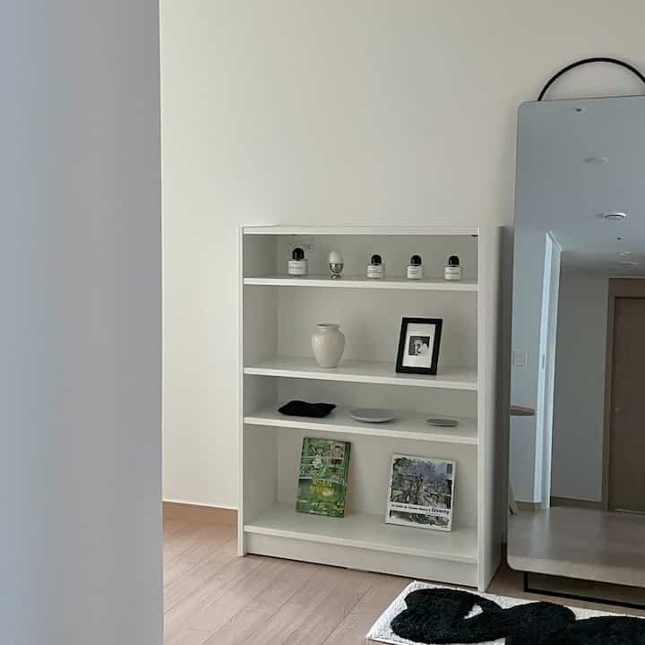 白い壁と本棚があるシンプルでナチュラルなリビングルーム、木目調の床と全身鏡が配置された落ち着いた雰囲気のインテリア