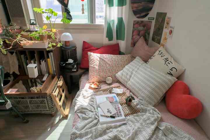 自然な色調と赤いクッションが配置された小さな寝室、窓辺に植物と水玉模様のカーテンがある雰囲気のよい空間