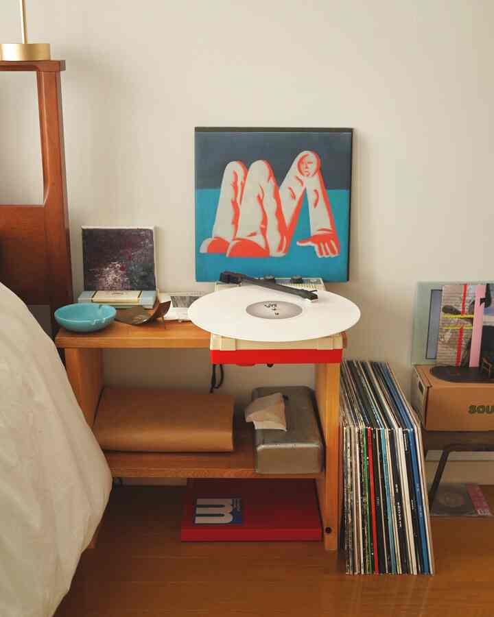 白い壁とブラウンの木製棚がある寝室で、ベッド横のDIY棚にレコードプレーヤーと様々な小物が配置された雰囲気のある空間