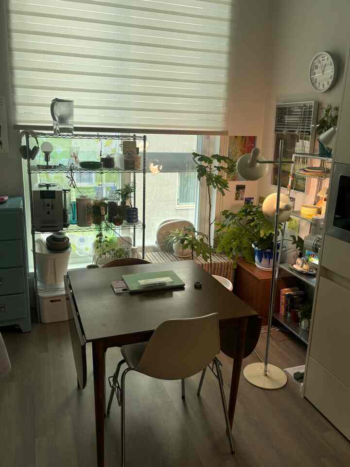 自然光と緑の植物が調和するダイニング、木製のテーブルと白い椅子、金属製のシェルビングユニットが配置された居心地の良いおうちカフェ空間