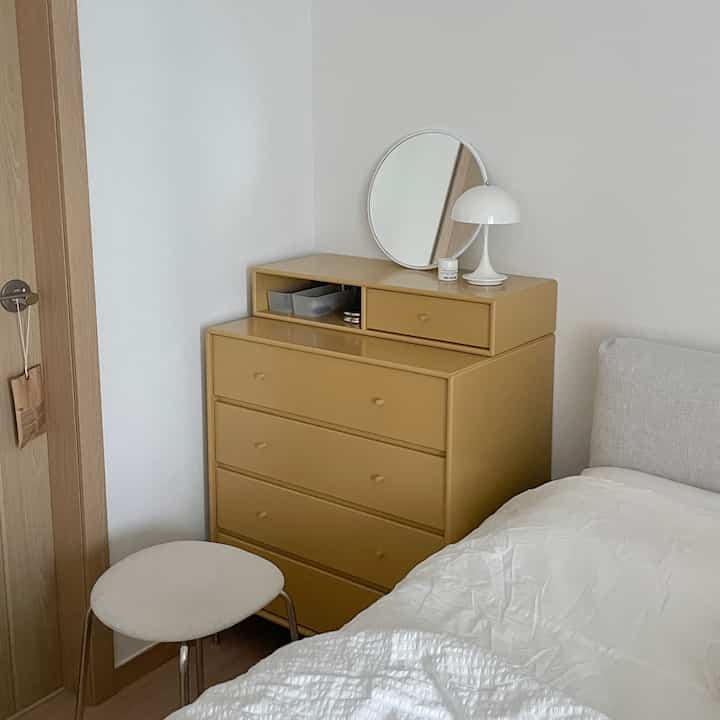 白い壁と寝具、ベージュのチェスト、丸い鏡とスツールが配置されたシンプルな寝室のインテリア