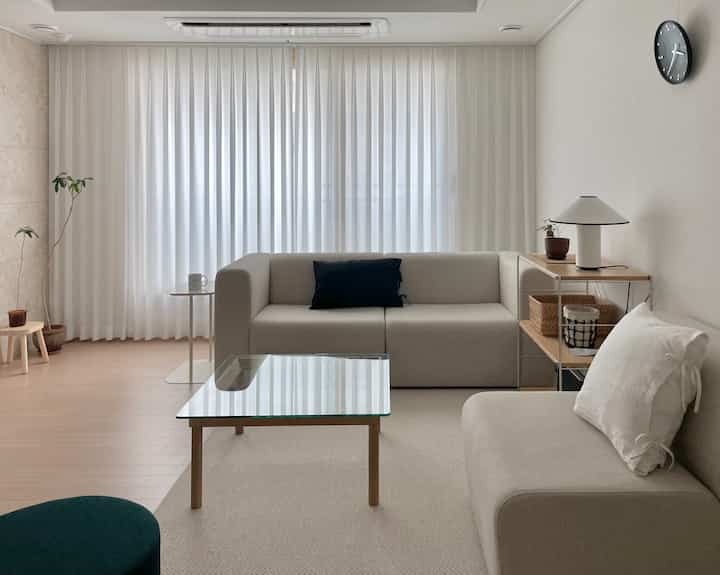 ナチュラルなベージュとホワイトの色調のリビング、2つのベージュソファとガラストップのコーヒーテーブル、白いカーテンが配置された清潔感のある空間