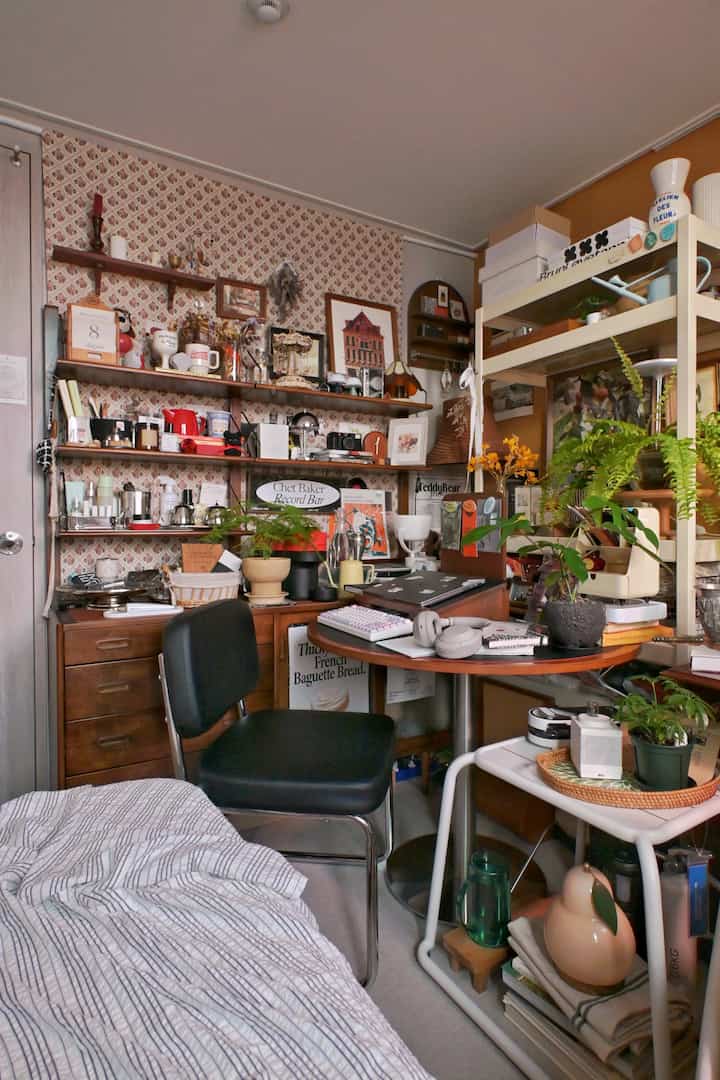 ナチュラルトーンの小さな寝室、デスクや棚に多様な小物や植物が配置された居心地の良いホームオフィス兼ホームカフェの空間