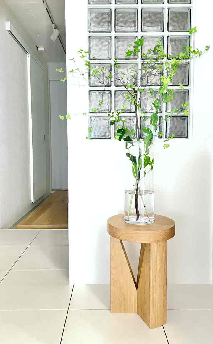 白いタイル床とガラスブロックの壁がある玄関、木目調のスツールの上に透明なガラス花瓶と生き生きとした緑の葉が置かれたミニマルな雰囲気のインテリア