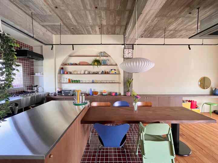 白い壁と天井が調和したキッチン兼ダイニングルーム、ステンレスのキッチンアイランドと木製ダイニングテーブル、多彩な椅子が配置されたナチュラルモダンなインテリア