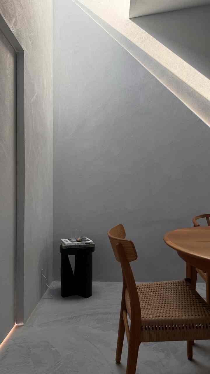 グレーの壁と床にナチュラルな木目調の丸いダイニングテーブルと椅子、黒いスツールが置かれたシンプルで自然なリビング空間